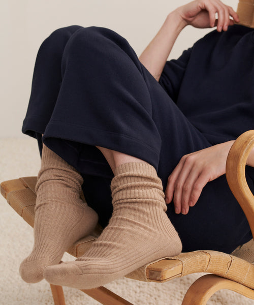 A person sits on a woven chair, legs crossed, wearing loose navy pants and Cashmere Socks Rewards GWP in caramel. Their upper body is partly visible; one hand touches their face while the other rests casually on their knee.