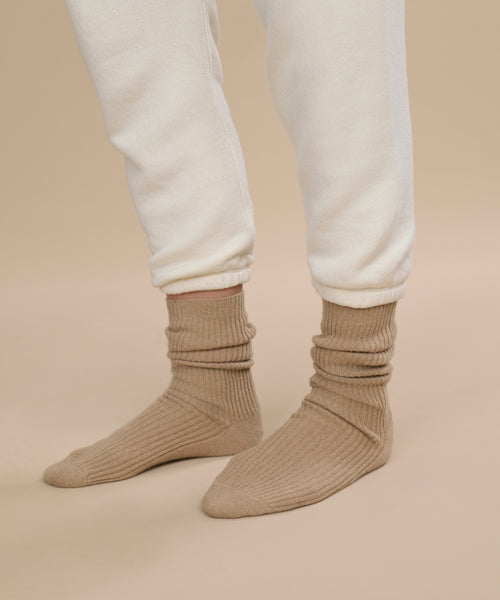 Model showcases caramel beige Cashmere Socks paired with off-white jogger pants against a beige floor and background, highlighting only the lower legs and feet for a cozy autumn/winter mood.