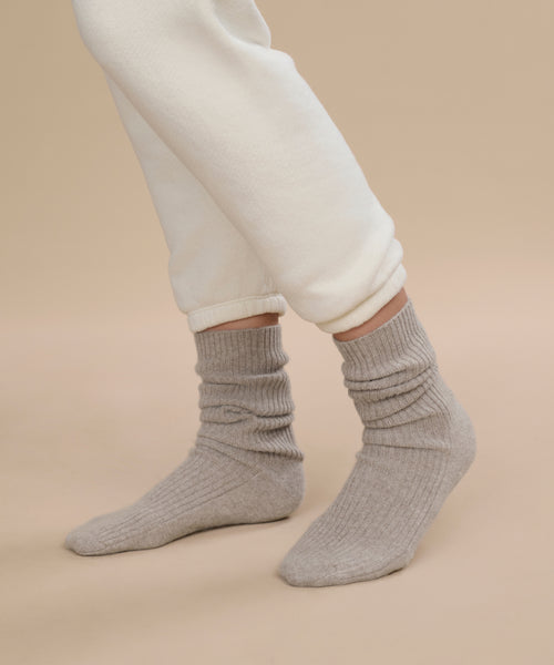 A person wearing off-white pants and cozy light gray ribbed Cashmere Socks stands on a neutral beige background.