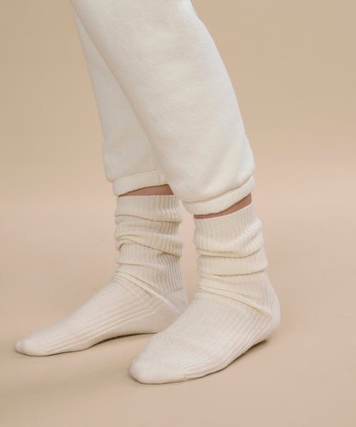 A person wears ivory Cashmere Socks, ideal for fall and winter, paired with off-white sweatpants. Only the lower legs and feet are visible against a light beige surface and neutral beige background.
