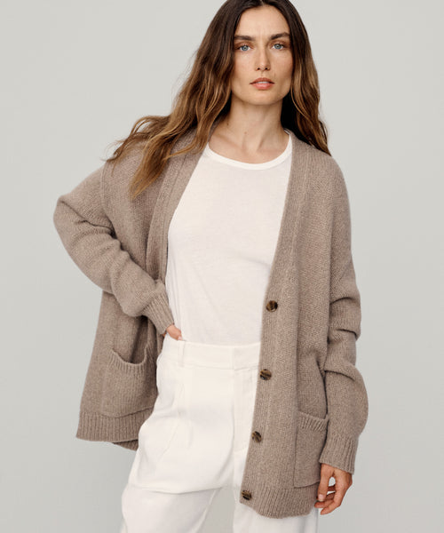 A woman with long brown hair models the Cashmere Spencer Cardigan in beige, layered over a white t-shirt and high-waisted white pants. She poses with one hand in her pocket against a plain light background.