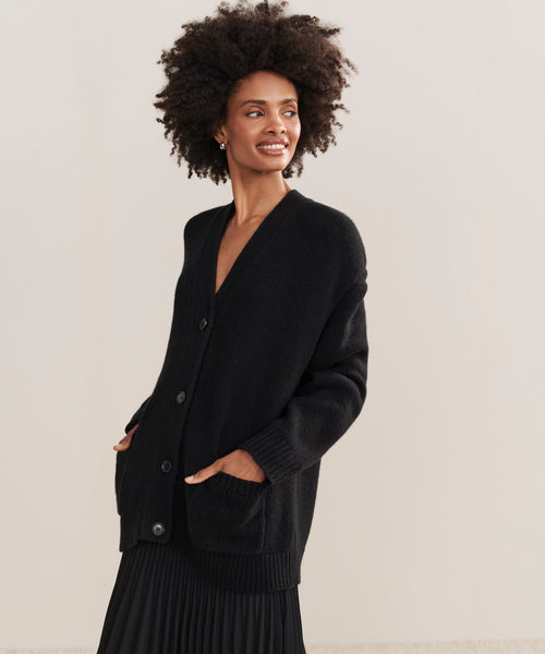 A woman with curly hair smiles, wearing the Cashmere Spencer Cardigan in a long black boyfriend fit, hands in the pockets, paired with a black pleated skirt, standing against a plain light-colored background.
