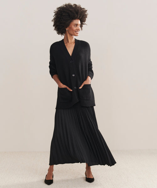 A woman with curly hair smiles while wearing a black Cashmere Spencer Cardigan, a black pleated skirt, and black flats. She stands with her hands in her pockets against a plain light background.