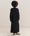 A person stands facing away, wearing a loose black Cashmere Spencer Cardigan in a boyfriend fit, a long black pleated skirt, and black pointed shoes with ankle straps. The background is neutral and minimal.