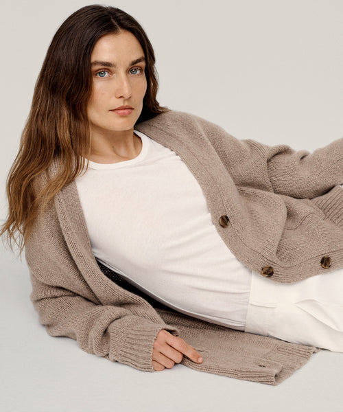 A woman with long brown hair reclines on her side against a neutral background, looking at the camera, wearing the Cashmere Spencer Cardigan over a white t-shirt and white pants.