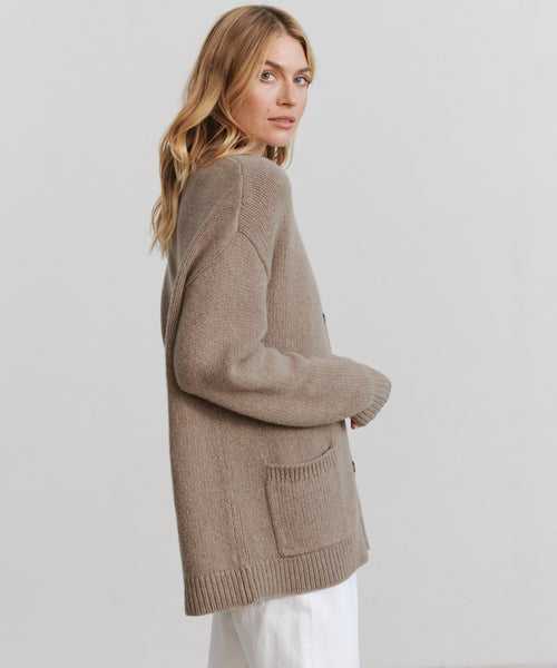 A blonde woman wears the Cashmere Spencer Cardigan in beige, featuring a cozy relaxed boyfriend fit, paired with white pants. She stands sideways against a plain light background.
