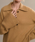 A person wears the Cashmere Stevie Polo, a camel knit top with large ribbed sleeves and an oversized collar, holding the sweater near the collar to display several gold rings. The persons face is partially out of frame.