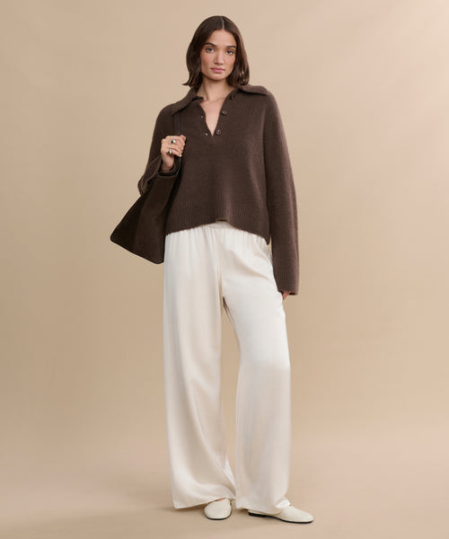 A woman stands against a beige background wearing the Cashmere Stevie Polo, wide-legged white pants, white shoes, and carrying a brown shoulder bag.