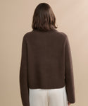 A woman with shoulder-length brown hair is shown from behind, wearing the Cashmere Stevie Polo and white pants, standing against a plain beige background.