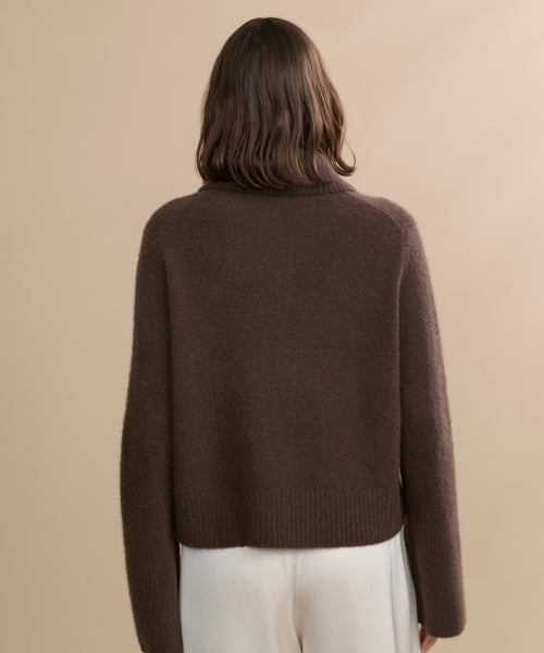 A woman with shoulder-length brown hair is shown from behind, wearing the Cashmere Stevie Polo and white pants, standing against a plain beige background.