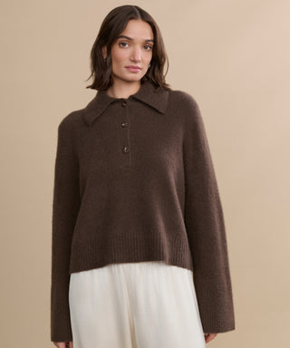 A woman with medium-length brown hair wears the Cashmere Stevie Polo, featuring an oversized collar and buttons, paired with loose white pants against a beige background.