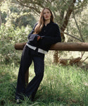 A woman with long hair leans against a wooden fence in a sunlit, grassy area, wearing the Cashmere Stevie Polo over loose black pants and a striped shirt, with trees and greenery in the background.
