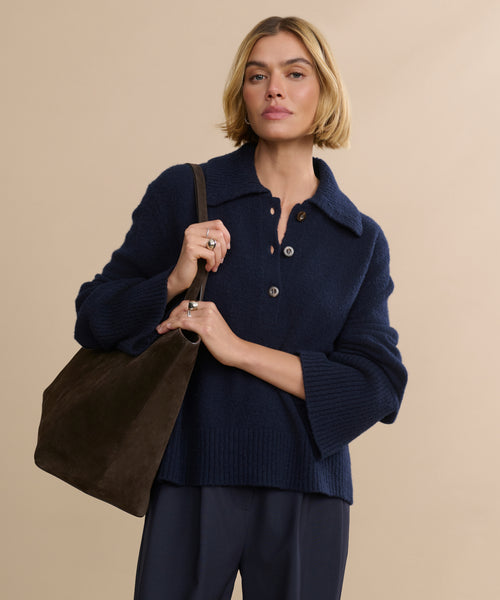 A woman with short blonde hair wears the Cashmere Stevie Polo in navy and dark trousers, carrying a large brown suede shoulder bag. She stands against a plain beige background, looking confidently at the camera.