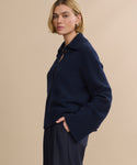 A woman with short blonde hair wears the Cashmere Stevie Polo—a navy sweater with an oversized collar and wide sleeves—paired with dark trousers, standing against a plain beige background.