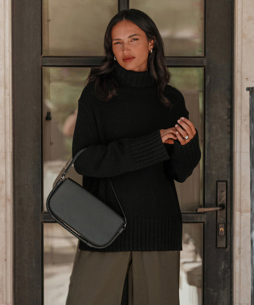 A woman with long dark hair wears the Cashmere Jet Turtleneck in black and olive green pants, standing by a glass door. She carries a black handbag over her shoulder and gazes calmly at the camera.