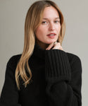 A woman with long blonde hair wears the oversized Cashmere Jet Turtleneck and a ring, gazing at the camera with a neutral expression against a plain background.