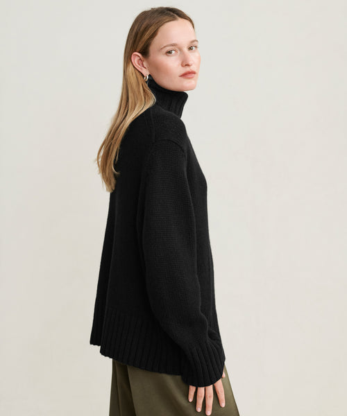 A woman with long, straight blonde hair models the oversized Cashmere Jet Turtleneck with olive green pants, standing sideways and looking over her shoulder against a plain backdrop.