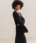 A woman with natural curly hair wears the Cashmere Spencer Cardigan in a boyfriend fit over a pleated black skirt, standing sideways against a neutral background and looking confidently at the camera.