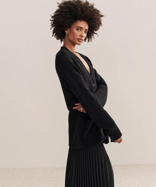 A woman with natural curly hair wears the Cashmere Spencer Cardigan in a boyfriend fit over a pleated black skirt, standing sideways against a neutral background and looking confidently at the camera.
