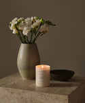 A Cedar Glass Candle, hand-poured in Los Angeles, sits lit on a stone surface beside a green ceramic vase with white flowers and a small dark bowl, all against a neutral beige background.