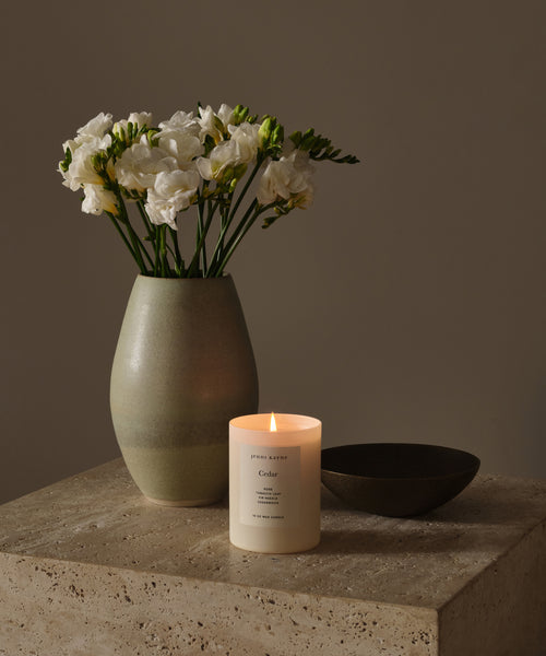 A Cedar Glass Candle, hand-poured in Los Angeles, sits lit on a stone surface beside a green ceramic vase with white flowers and a small dark bowl, all against a neutral beige background.