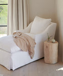 A cozy white chaise lounge with a beige throw sits beside the Cedar Stump, a small round side table crafted from reclaimed cedarwood, which holds a vase of flowers in a bright room with light wood floors and cream curtains.