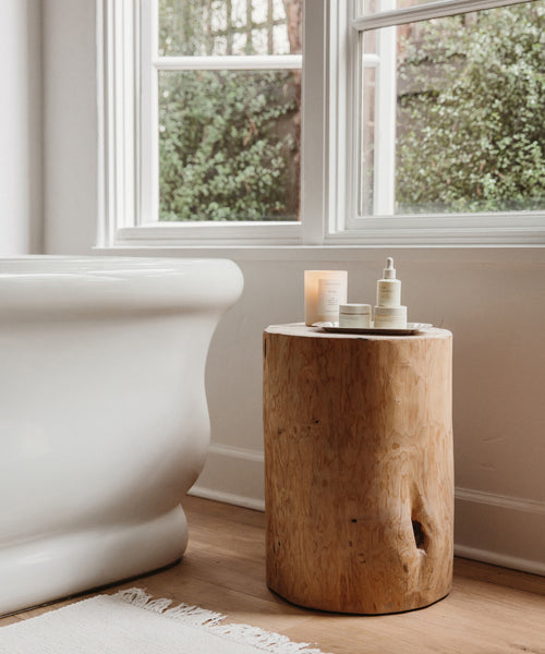 A white bathtub sits next to the Cedar Stump side table—crafted from reclaimed cedarwood—holding skincare bottles and a candle, in a bright room with large windows framing lush greenery for a perfect natural accent.
