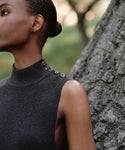 A woman in the Celia Sweater Dress—sleeveless with a mock neck and shoulder buttons in dark gray—stands beside a tree with her hair pulled back, greenery softly blurred in the background.