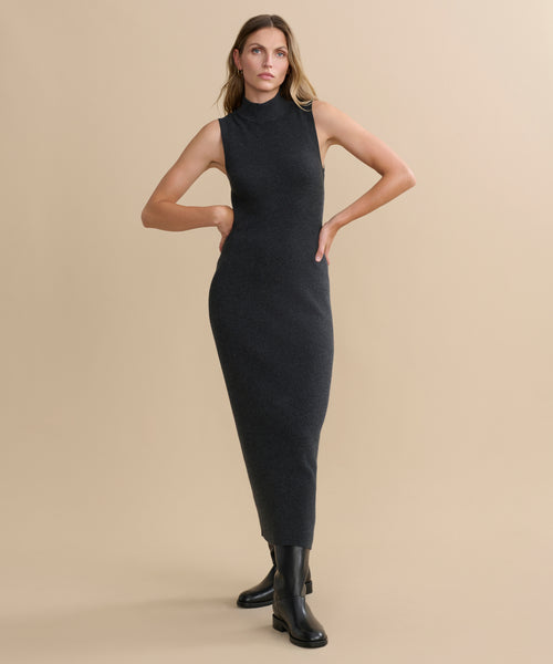 A woman with straight, loose hair stands against a beige background wearing the Celia Sweater Dress, a sleeveless, fitted dark gray midi style in cotton cashmere, paired with black ankle boots and her hands on her hips.