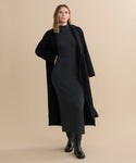 A woman stands against a beige background wearing the fitted, ankle-length Celia Sweater Dress in dark grey cotton cashmere, layered under a long black coat with black boots. She has wavy, shoulder-length hair and looks slightly to the side.