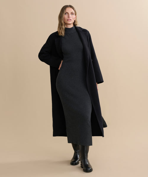 A woman stands against a beige background wearing the fitted, ankle-length Celia Sweater Dress in dark grey cotton cashmere, layered under a long black coat with black boots. She has wavy, shoulder-length hair and looks slightly to the side.