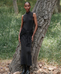 Wearing the Celia Sweater Dress and black boots, a woman stands outdoors leaning against a tree trunk, surrounded by green grass and scattered rocks.