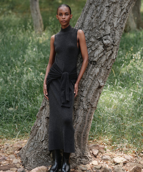 Wearing the Celia Sweater Dress and black boots, a woman stands outdoors leaning against a tree trunk, surrounded by green grass and scattered rocks.