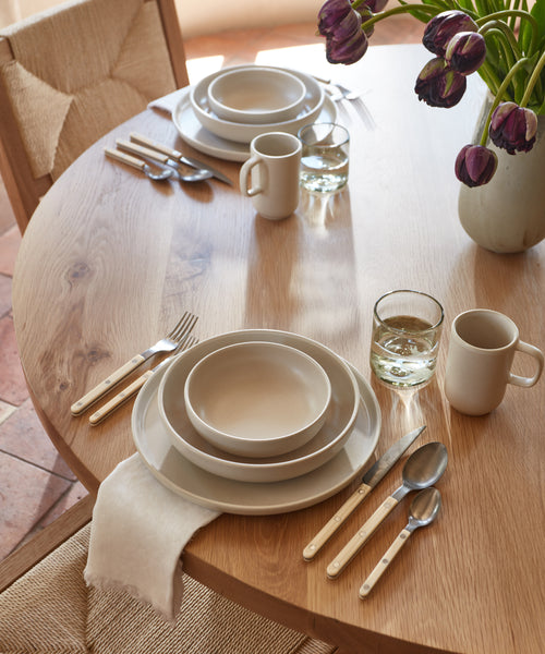 A wooden dining table set with neutral-toned plates, bowls, mugs, the Bistro Vintage Finish Cutlery Set with white handles, glasses of water, a white napkin, and a vase of purple tulips. Sunlight streams onto the table.