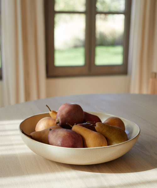 The Pacific Serving Bowl in beige, filled with pears, rests on a light wooden table as sunlight pours through sheer curtains, creating a cozy atmosphere.