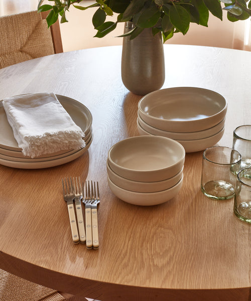 A wooden table set with stacked beige stoneware plates and Pacific Soup Bowls, four metal forks with white handles, three clear glasses, a white napkin, and a vase of green leafy branches.