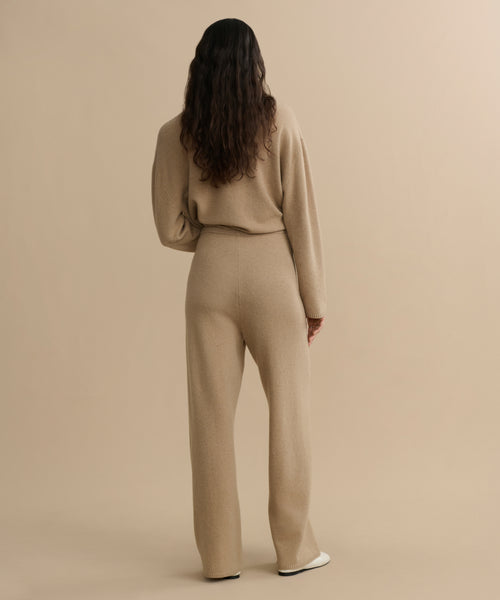 A person with long, wavy hair faces away, wearing the Chalet Pant— a loose-fitting, beige felted wool loungewear set—against a plain, light beige background.