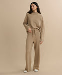 Woman with long dark hair wears the Chalet Pant, a loose-fitting, beige wide-leg knit loungewear set with a drawstring waist. She stands against a plain beige background, looking slightly to the side.