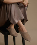 Person wearing brown suede mules sitting on a black stool against a beige background