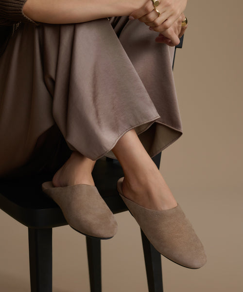 Person wearing brown suede mules sitting on a black stool against a beige background