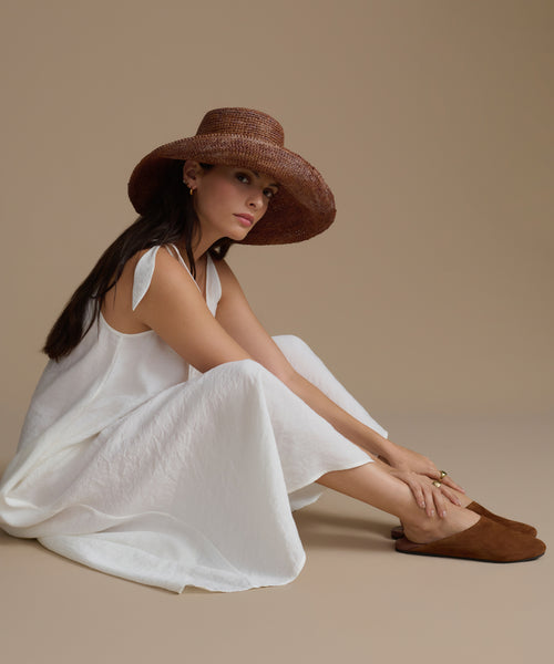 Woman wearing a white dress, brown sun hat and brown leather mules against a beige background.