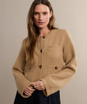 A woman with long brown hair models the Charlotte Cardigan, a beige knit camel sweater featuring large buttons and front pockets. She stands against a light background, gently smiling at the camera with her hands clasped in front.