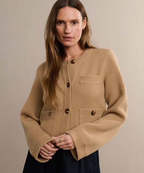 A woman with long brown hair models the Charlotte Cardigan, a beige knit camel sweater featuring large buttons and front pockets. She stands against a light background, gently smiling at the camera with her hands clasped in front.