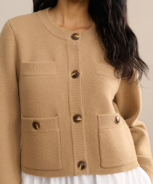 A woman models the Charlotte Cardigan, a tan button-up knit with four front pockets and brown buttons, stylishly layered over a white skirt or dress. The photo is cropped to her torso and part of her face.