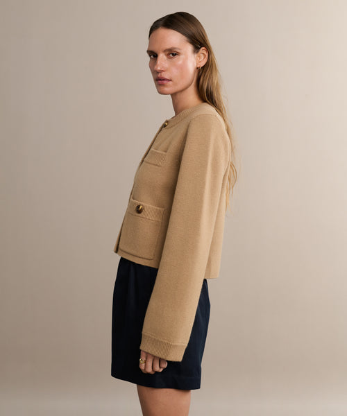 A woman with long, light brown hair stands sideways in a Charlotte Cardigan with front pockets and buttons, styled with dark shorts—an ideal women’s fashion choice—set against a plain beige background.