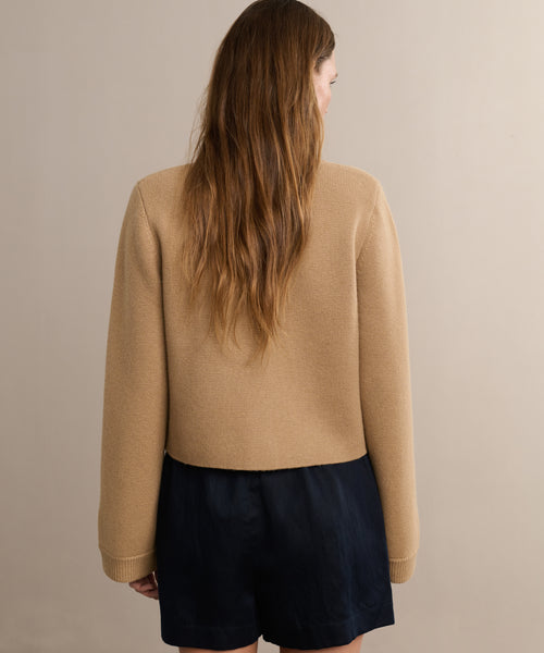 A woman with long wavy brown hair faces away from the camera, wearing the Charlotte Cardigan and black shorts, embodying modern womens style against a simple beige background.