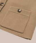 Close-up of the Charlotte Cardigan, a beige knit camel women’s cardigan featuring a ribbed square front pocket with a brown button, plus another ribbed edge and button visible on the left.