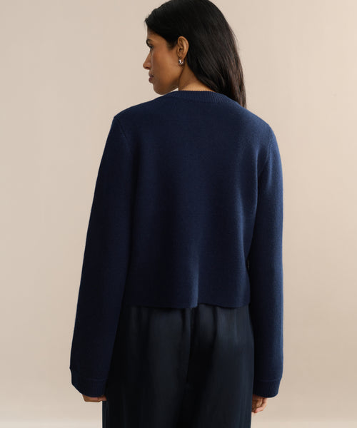 A woman with long dark hair faces away, wearing the Charlotte Cardigan in navy with long sleeves and black pants against a plain beige background.