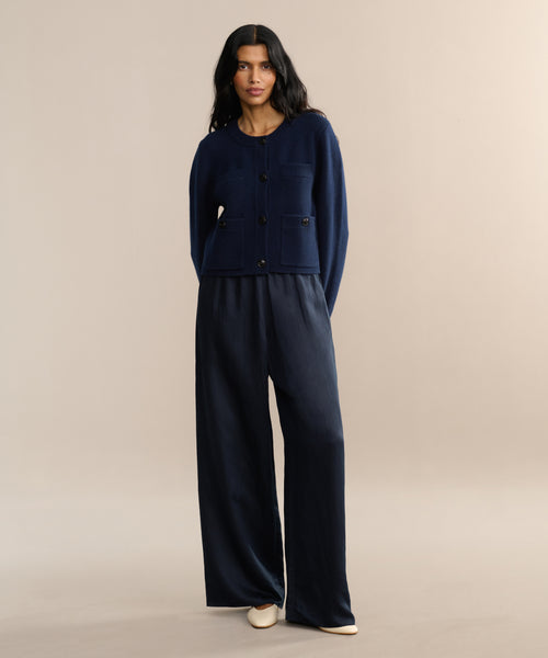 A woman with long dark hair models the Charlotte Cardigan, a navy button-up women’s cardigan with front pockets, paired with wide-leg navy pants and cream shoes. She stands facing forward against a neutral background.