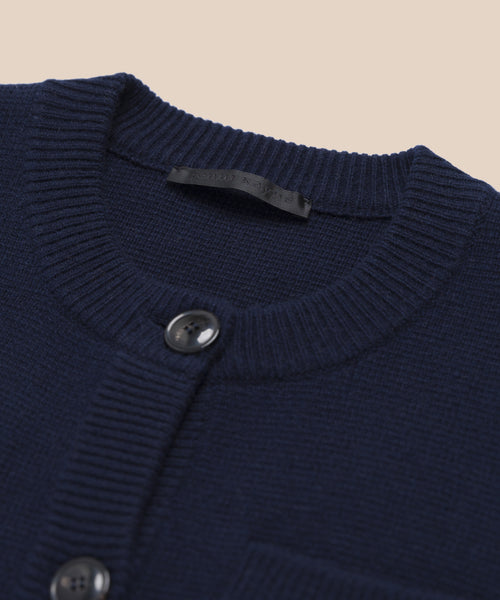 Close-up of the Charlotte Cardigan in navy blue, featuring a ribbed crew neckline, two black buttons, and a black label at the collar. This womens cardigan is displayed against a solid beige background.